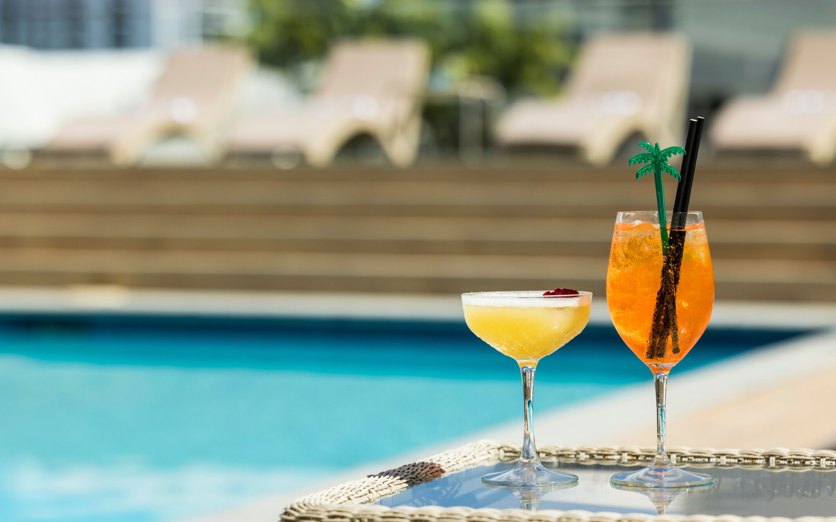 Two cocktails on a table by a poolside with lounge chairs in the background.