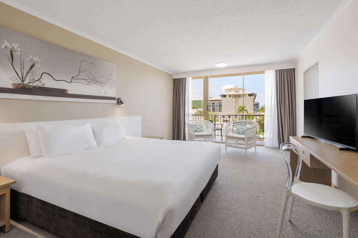 A hotel room with a large bed, a desk, a TV, and a balcony with chairs.