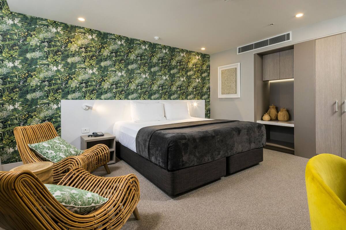 A modern bedroom with a large bed, a nightstand, a wicker chair, a yellow chair, and a wardrobe.