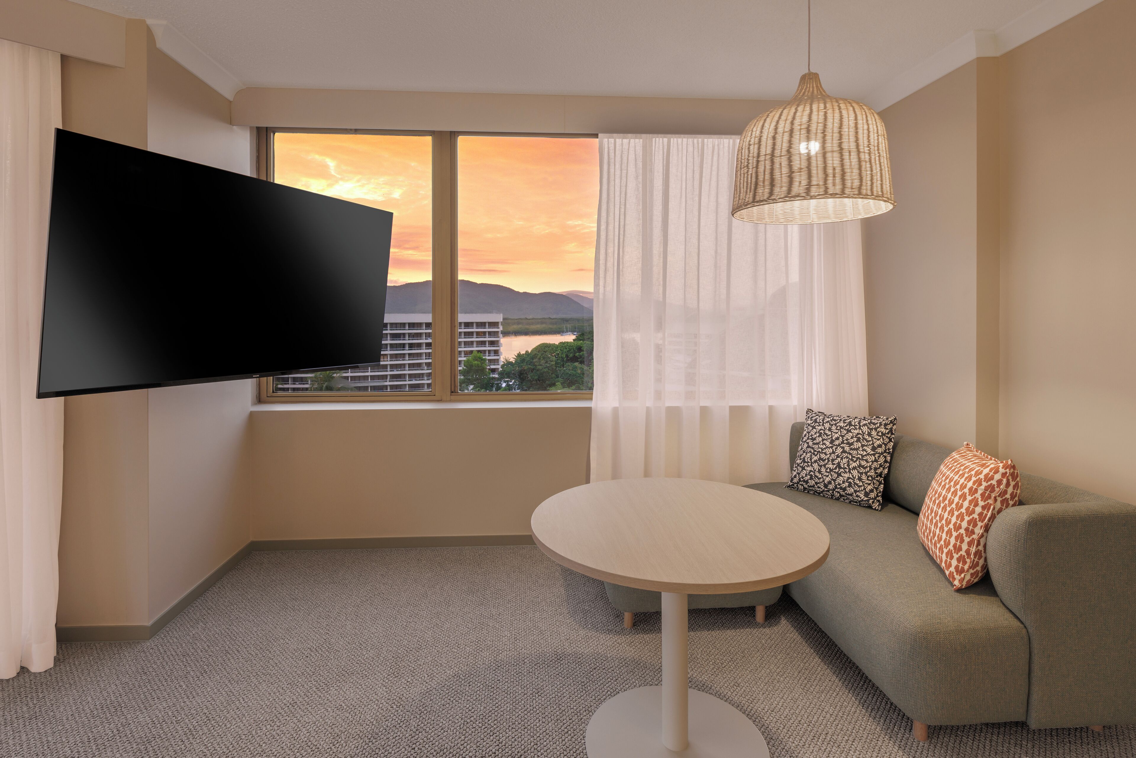 A modern hotel room with a flat-screen TV, a round table, a sofa with patterned cushions, and a view of a sunset over mountains.
