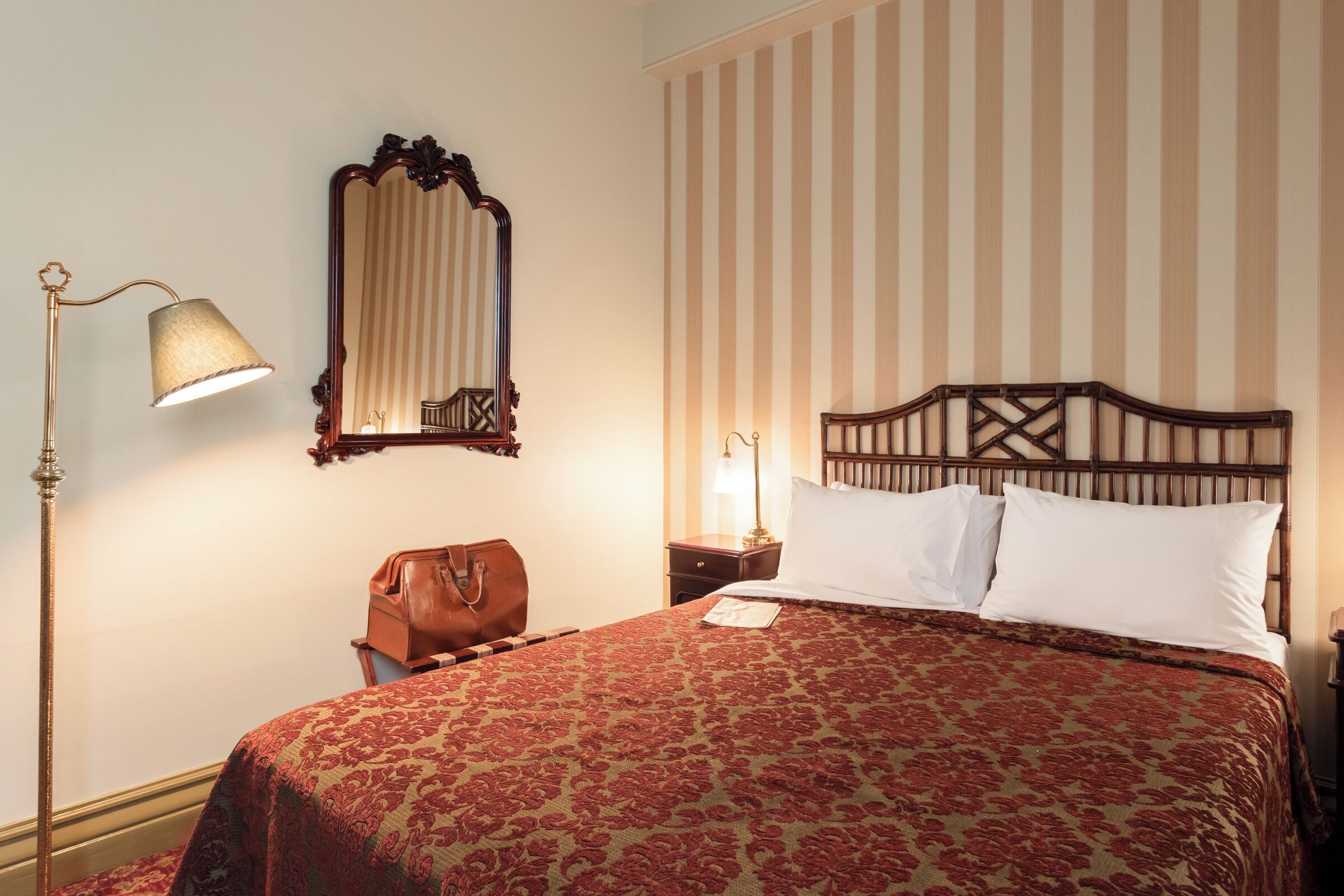 A neatly made bed with a patterned bedspread, a wooden headboard, a bedside lamp, a mirror, and a striped wall.