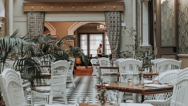Un comedor con sillas y mesas de mimbre blanco, suelo a cuadros y la vista de una persona al fondo.