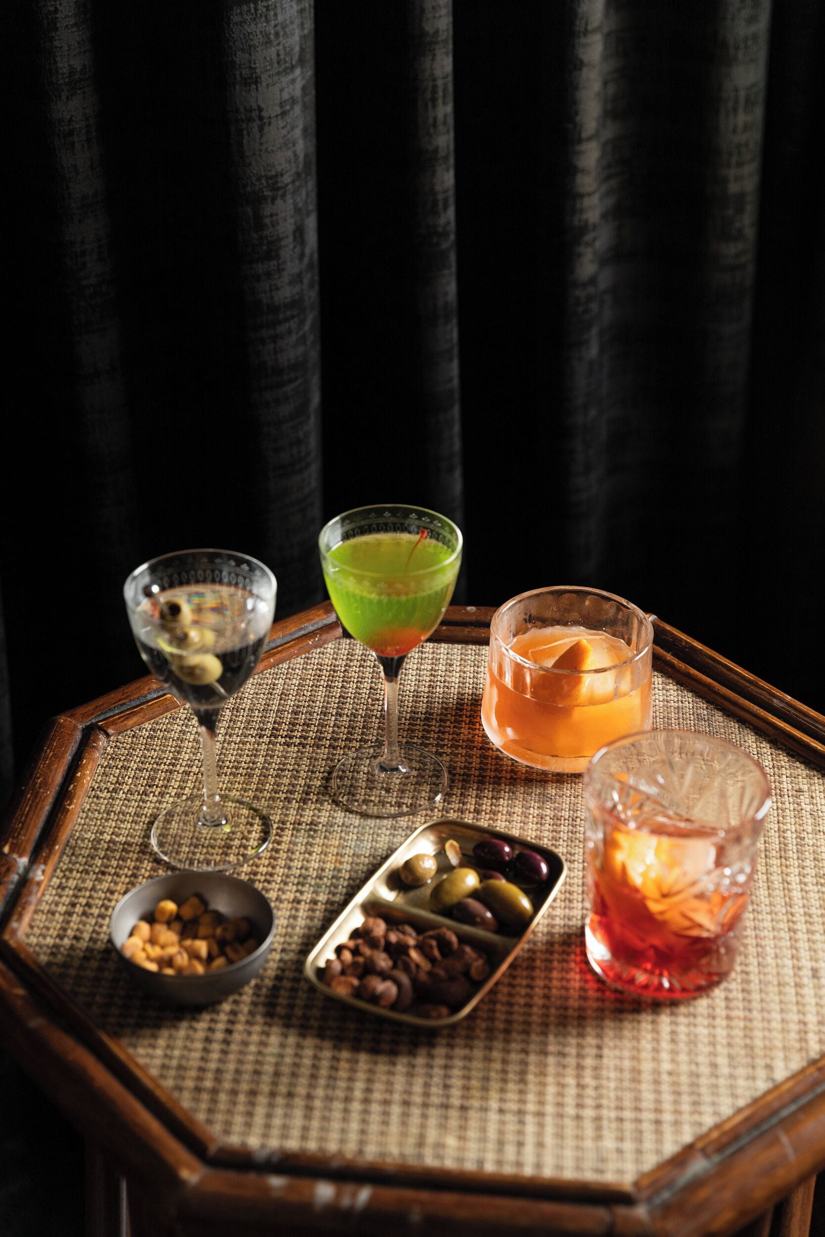 A table with three glasses of different colored drinks, a bowl of nuts, and a tray of olives.