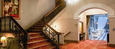 A grand staircase with a chandelier, a red carpeted floor, and a painting of a royal figure.