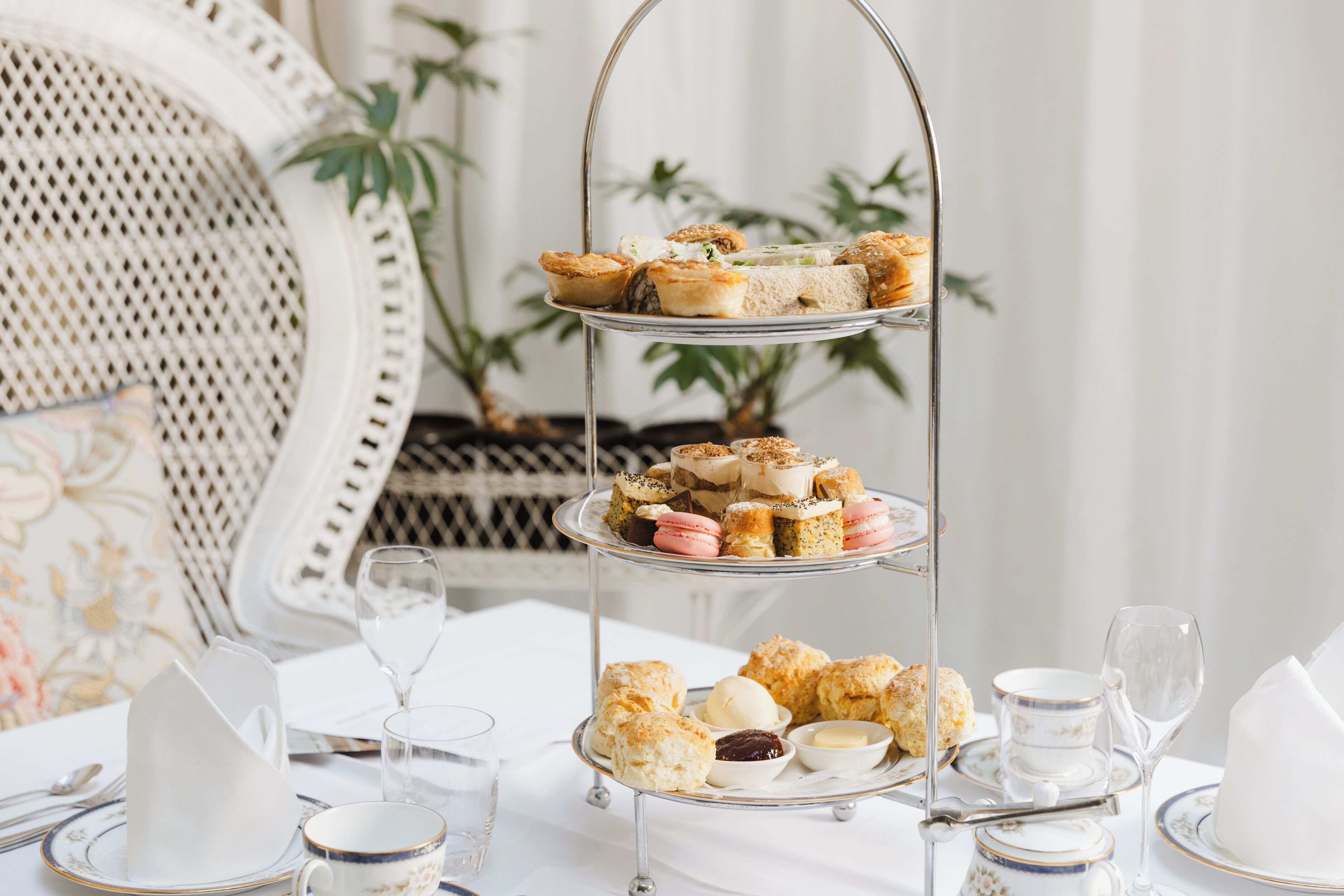 A three-tiered dessert stand with pastries and scones, surrounded by white tableware and a wicker chair.