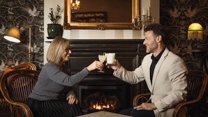 Una pareja brindando con bebidas en una habitación cálidamente iluminada con chimenea, muebles de mimbre y papel tapiz estampado.