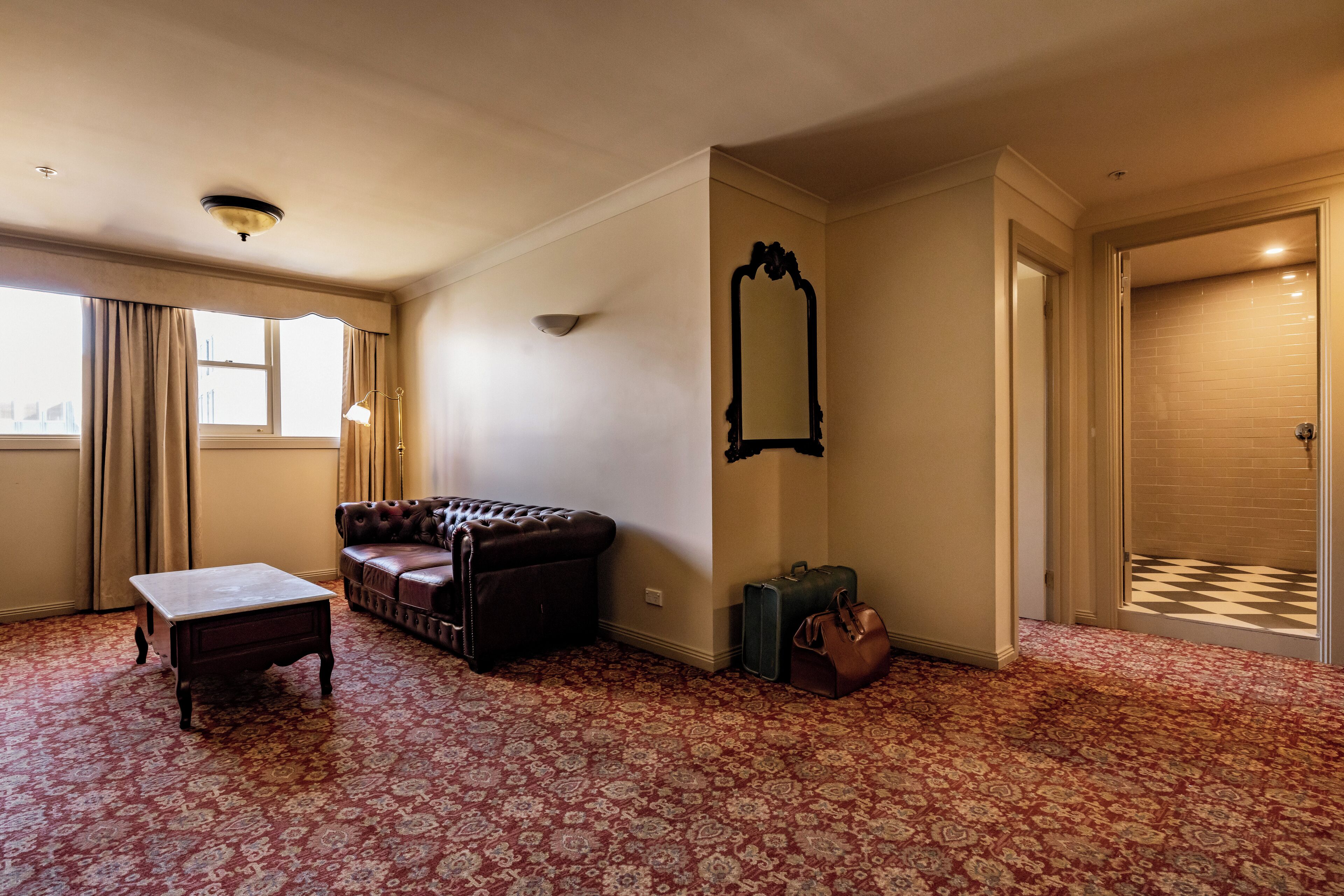 Una habitación con alfombra estampada, un sofá de cuero, una mesita, un espejo y un suelo a cuadros que conduce a una puerta.