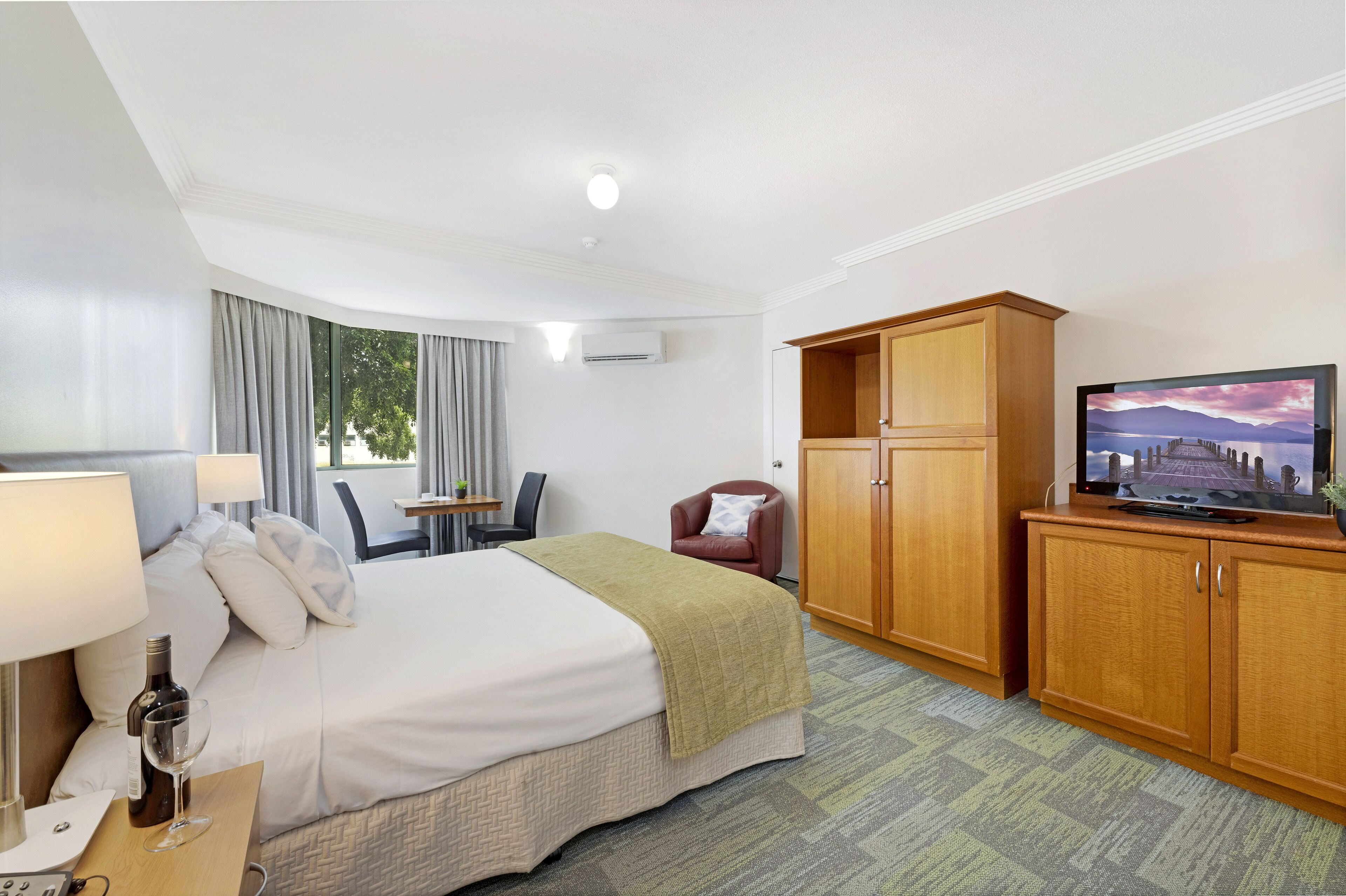 A hotel room with a bed, a television, a desk with a chair, a wooden wardrobe, and a window with curtains.