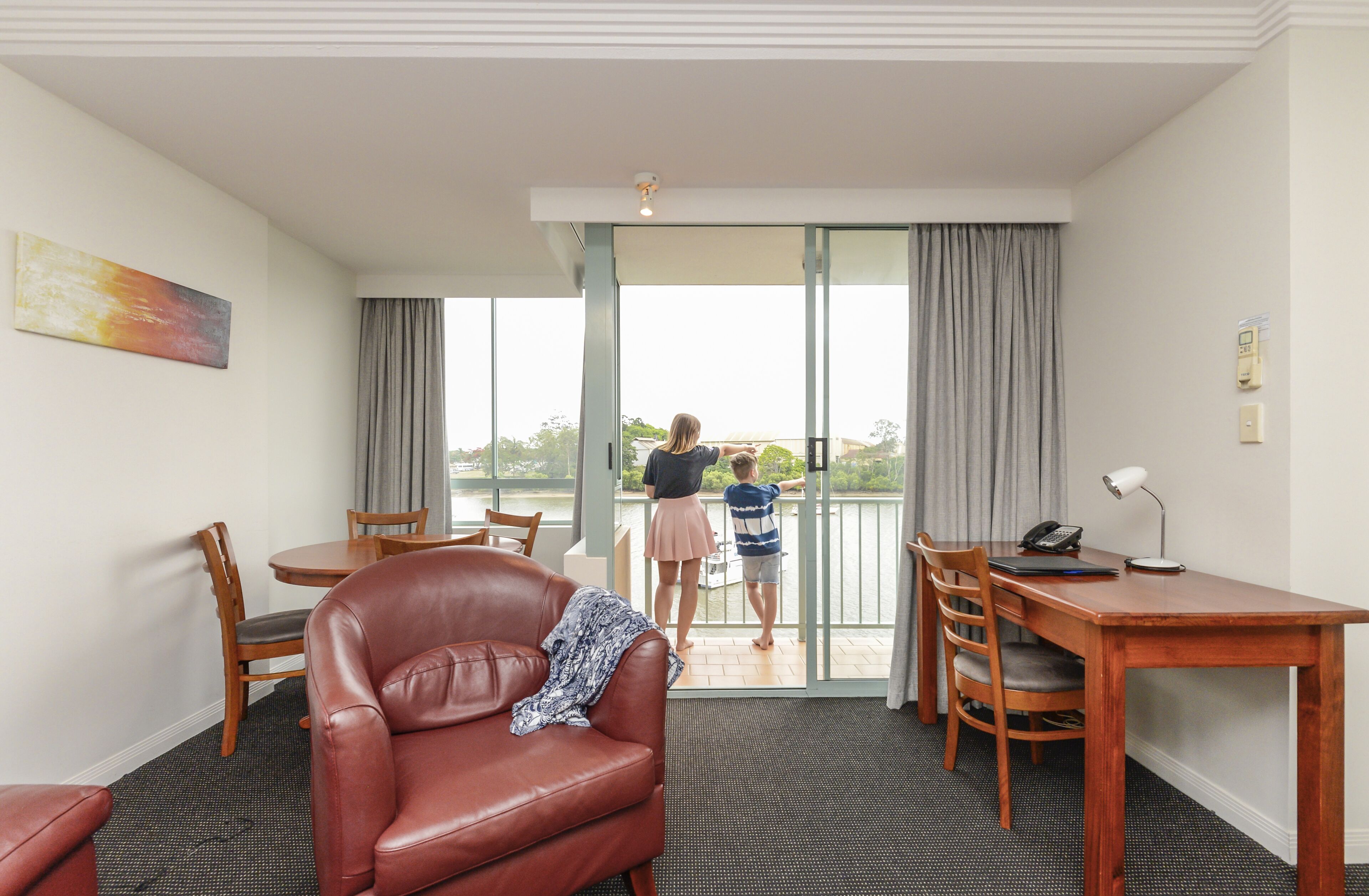 Habitación de hotel con comedor, una butaca de cuero, escritorio con lámpara y vista a un balcón con dos personas.