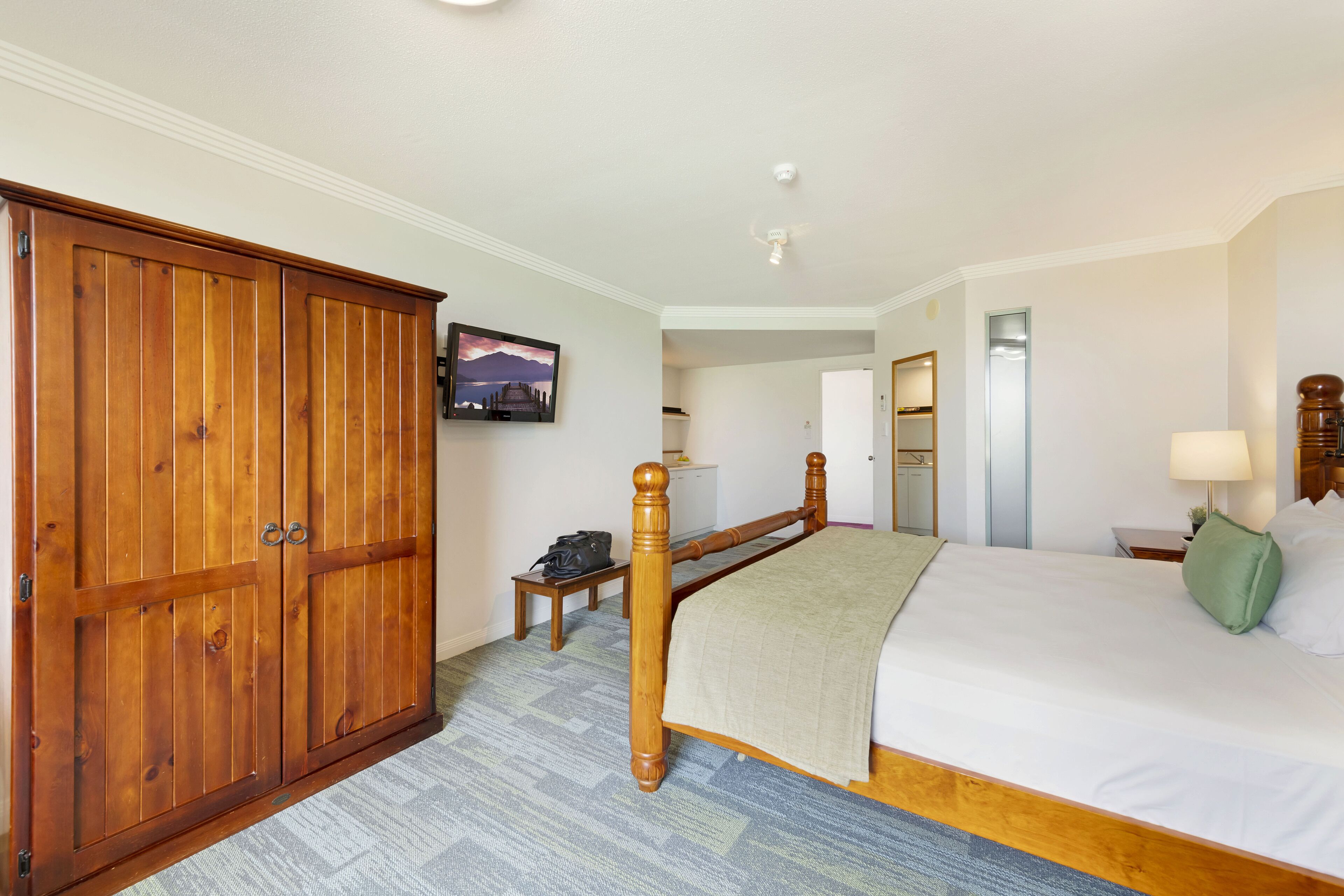 Ein Hotelzimmer mit großem Holzschrank, einem Bett mit weißer Bettwäsche, einem kleinen Nachttisch mit Lampe und einem an der Wand montierten Fernseher.