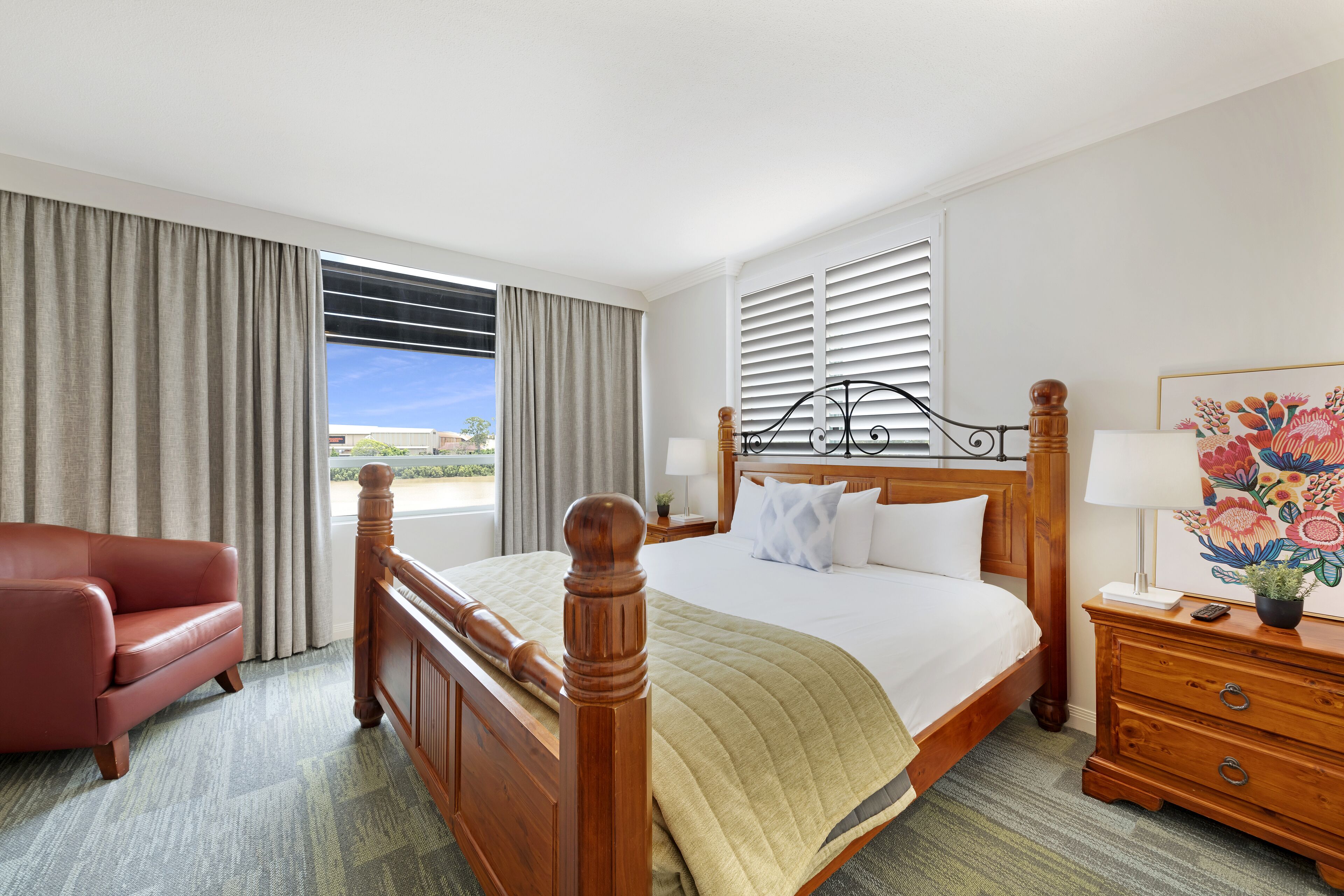 A hotel room with a large bed, a wooden headboard, a red armchair, a nightstand with a lamp, and a view of the outdoors.
