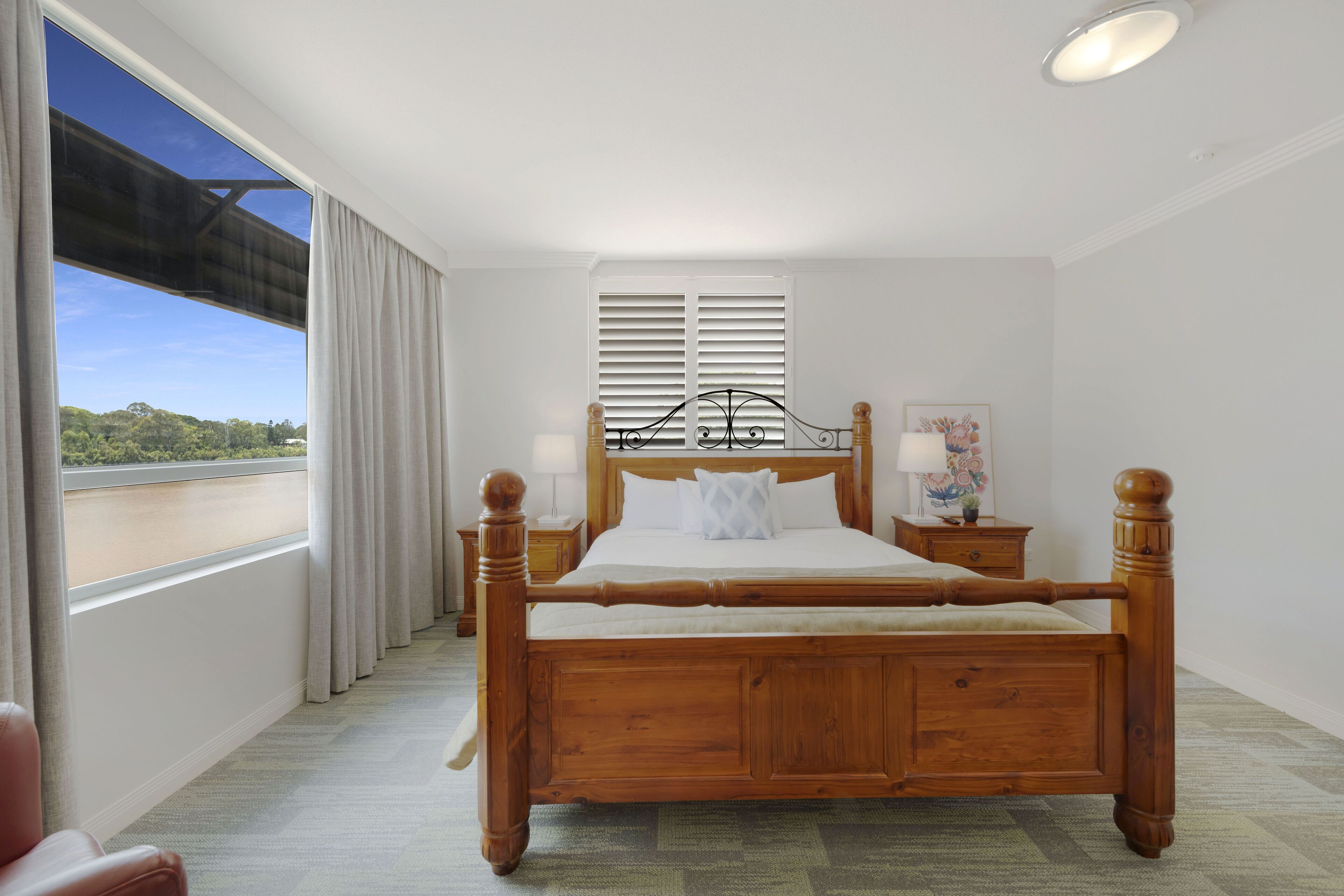 A wooden four-poster bed with white linens, a nightstand with a lamp, and a window with curtains.