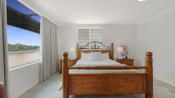 A wooden four-poster bed with white linens, a nightstand with a lamp, and a window with curtains.