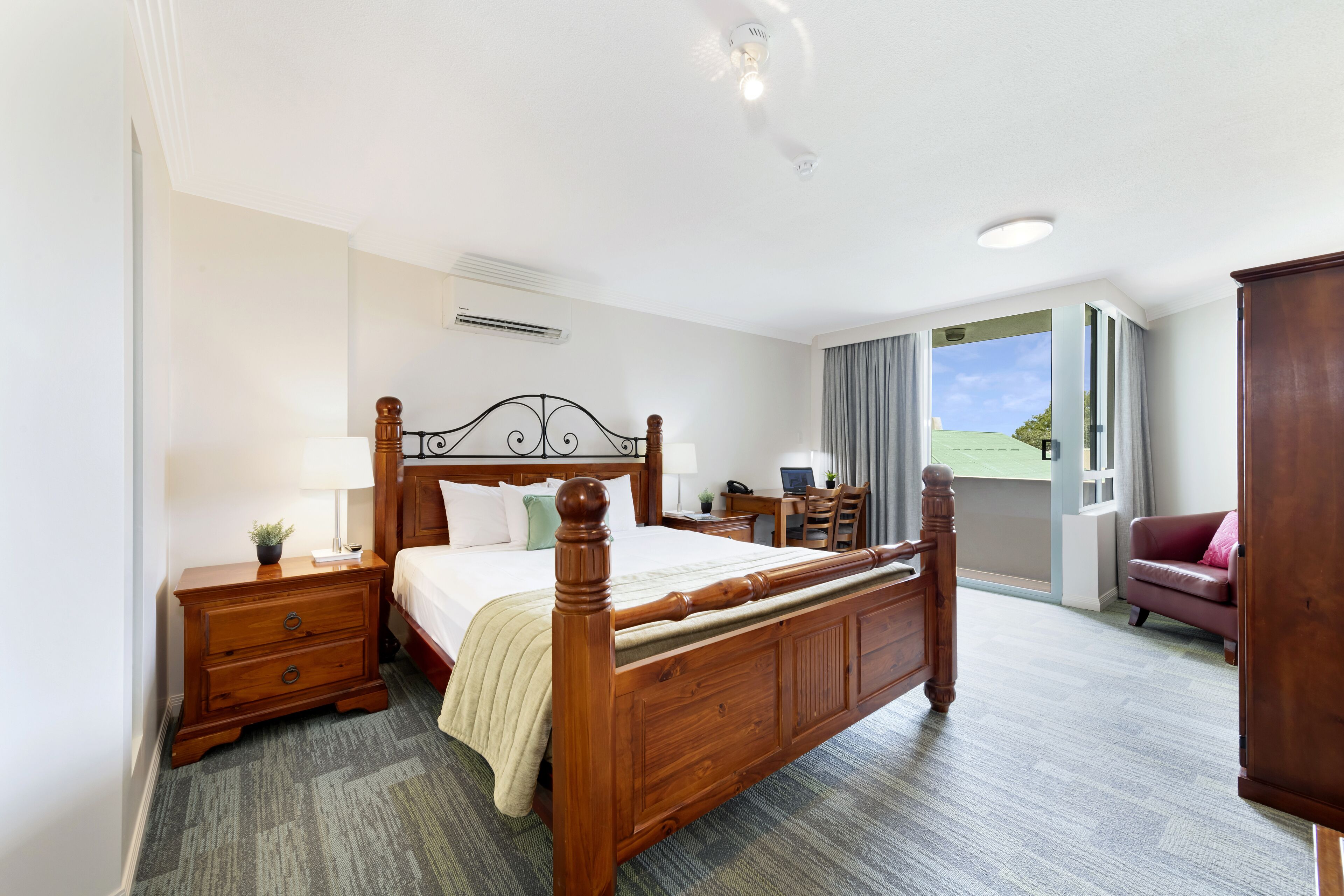 Une chambre d’hôtel avec un lit à baldaquin en bois, un bureau avec une chaise, un canapé et une grande fenêtre donnant sur l’extérieur.