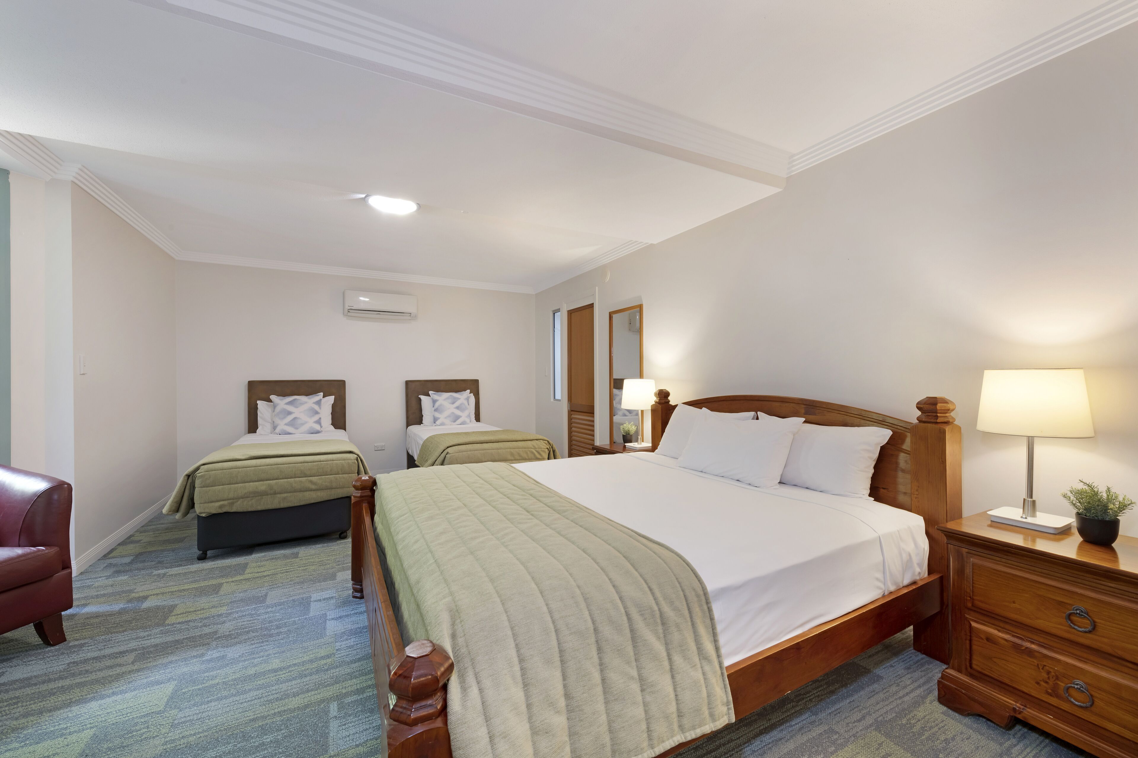 A hotel room with a large bed, two smaller beds, a wooden nightstand, a chair, and a carpeted floor.