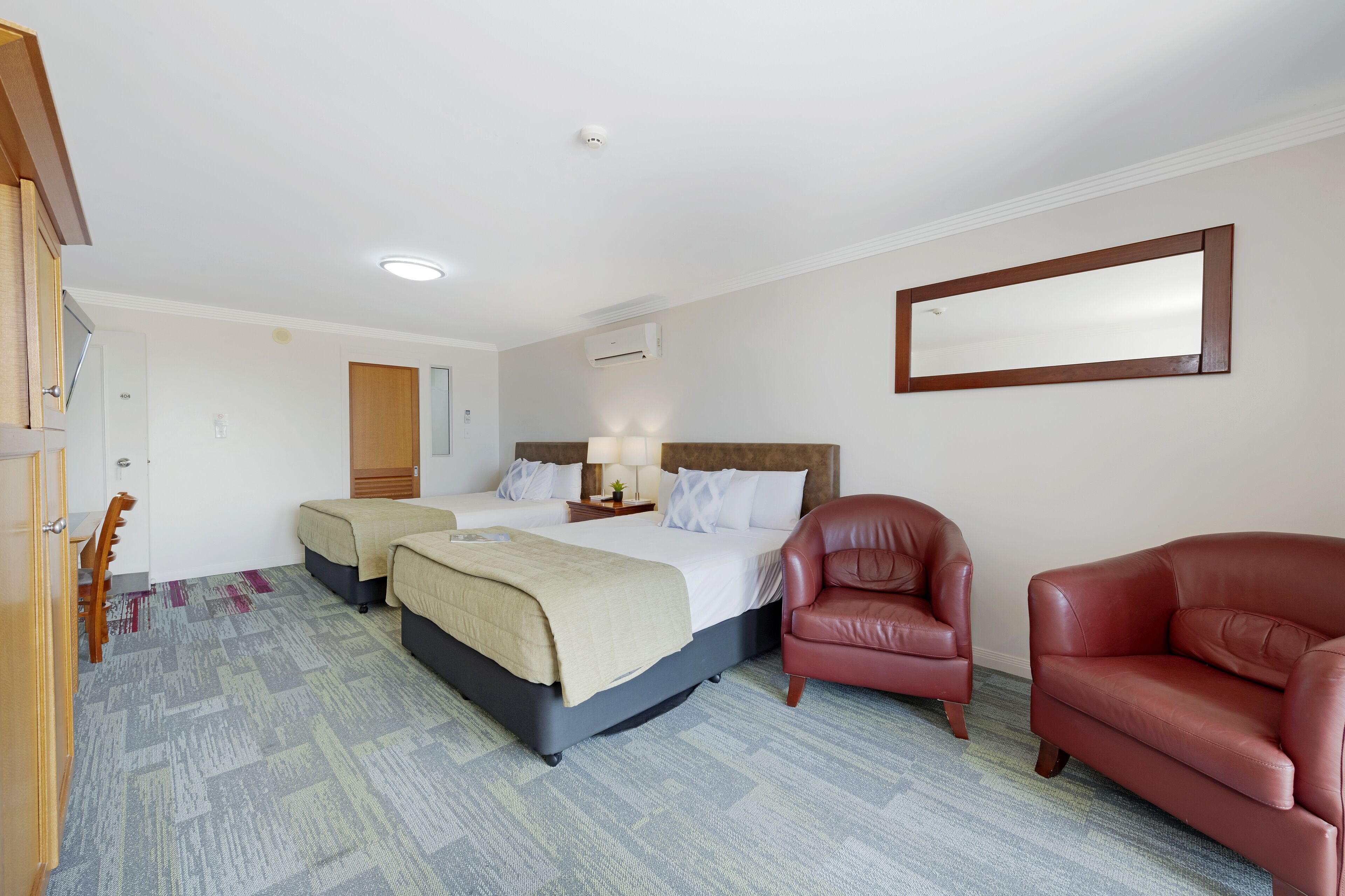 Une chambre d’hôtel avec deux lits, un fauteuil en cuir rouge, une armoire en bois et un miroir au mur.