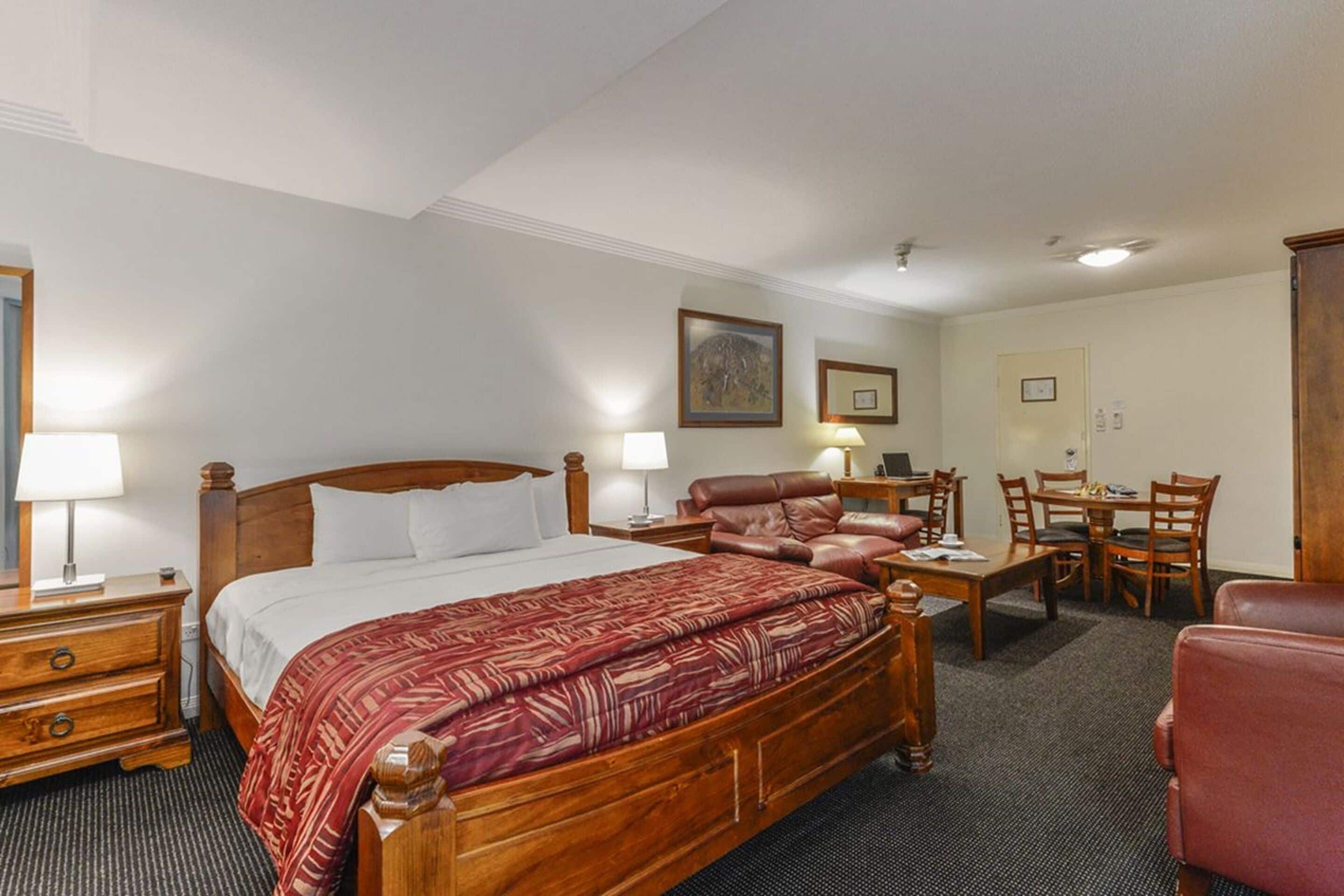 A hotel room with a wooden bed, a leather sofa, a dining table with chairs, and a desk with a chair.