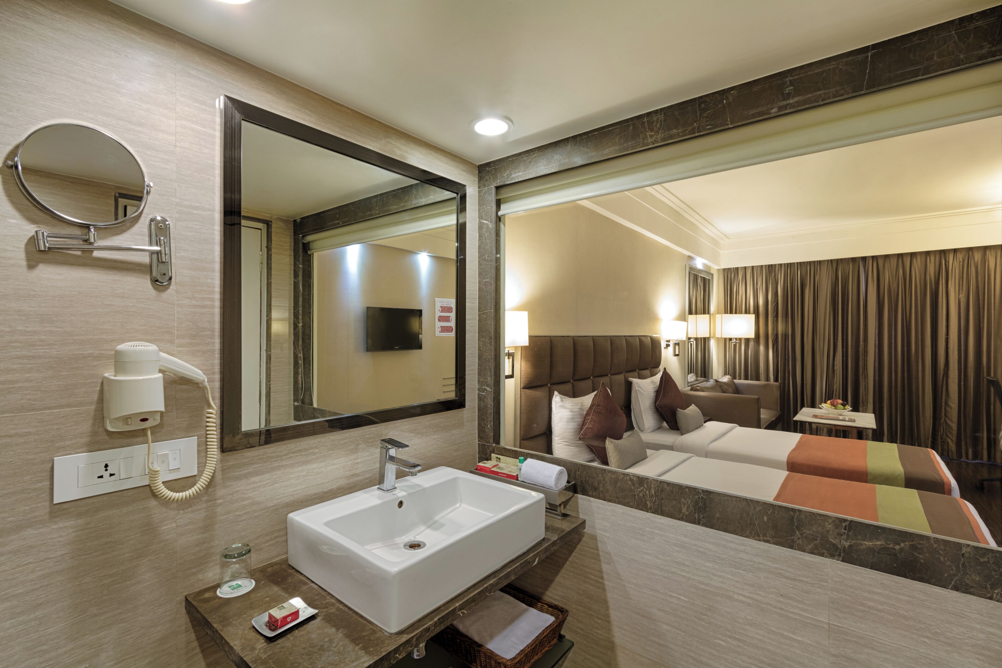 A hotel bathroom with a sink, mirror, hairdryer, and a bed with a view of the room.