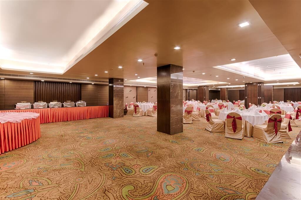 A spacious banquet hall with a patterned carpet, rows of tables with white tablecloths and gold sashes, and a high ceiling with recessed lighting.