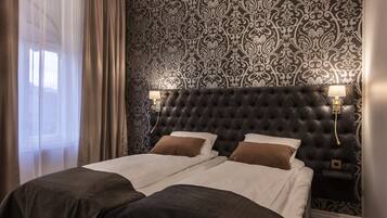 Habitación de hotel con cabecera capitonada, dos camas con ropa de cama blanca y marrón, un papel tapiz estampado y dos lámparas de noche.