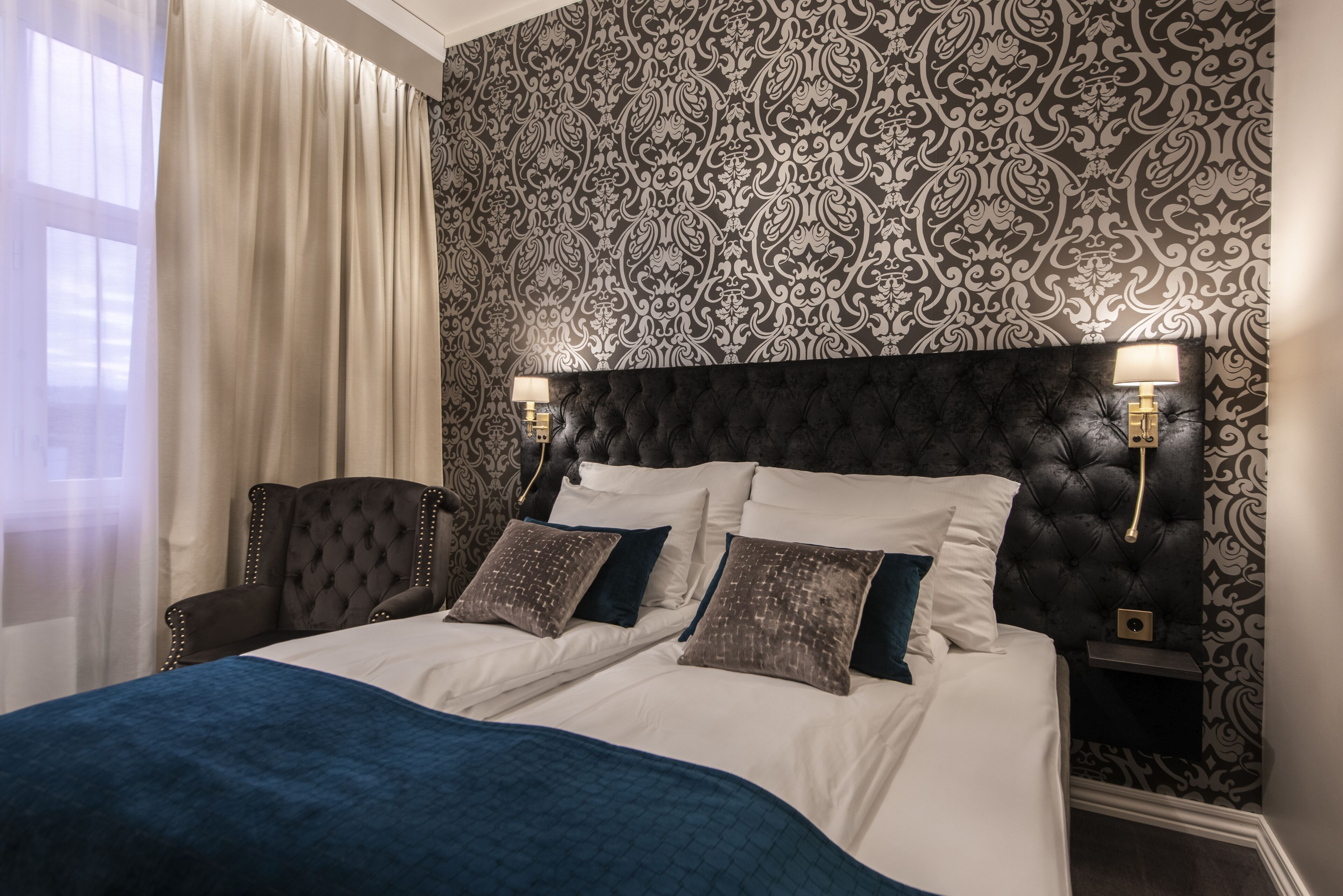 A bedroom with a tufted headboard, two bedside lamps, a large bed with a blue blanket, and a patterned wallpaper.