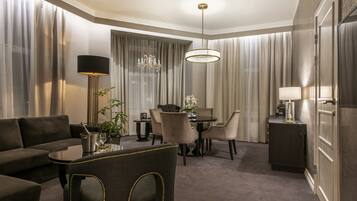 A hotel room with a dark-colored sofa, a dining table with chairs, a chandelier, and a cabinet with a lamp.