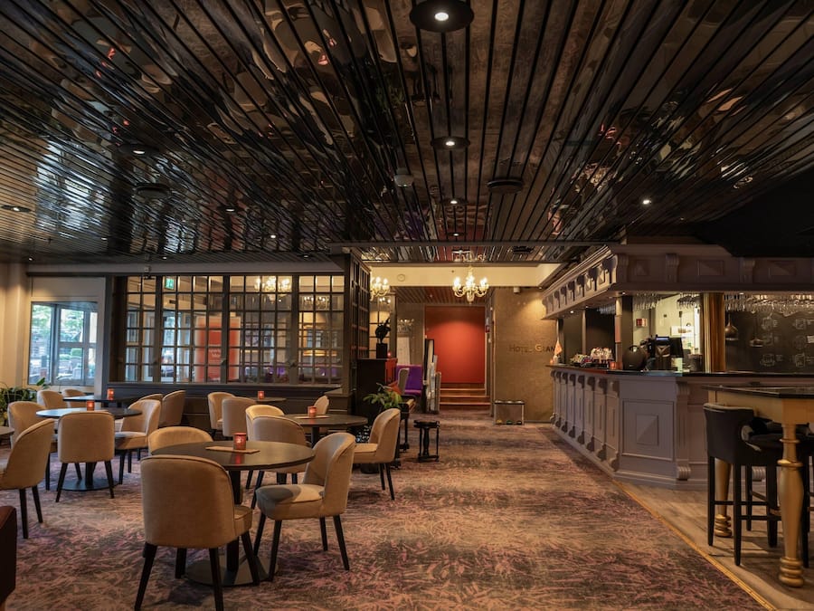 Un'accogliente hall d'albergo con tavoli rotondi, sedie, un'area bar e una scala che conduce ai piani superiori.