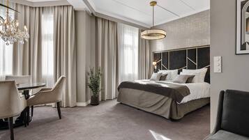 A modern hotel room with a large bed, a dining area with chairs, a chandelier, and a large window with curtains.