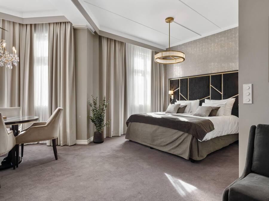 Una habitación de hotel moderna con una cama grande, una zona de comedor con sillas, una araña y un ventanal con cortinas.