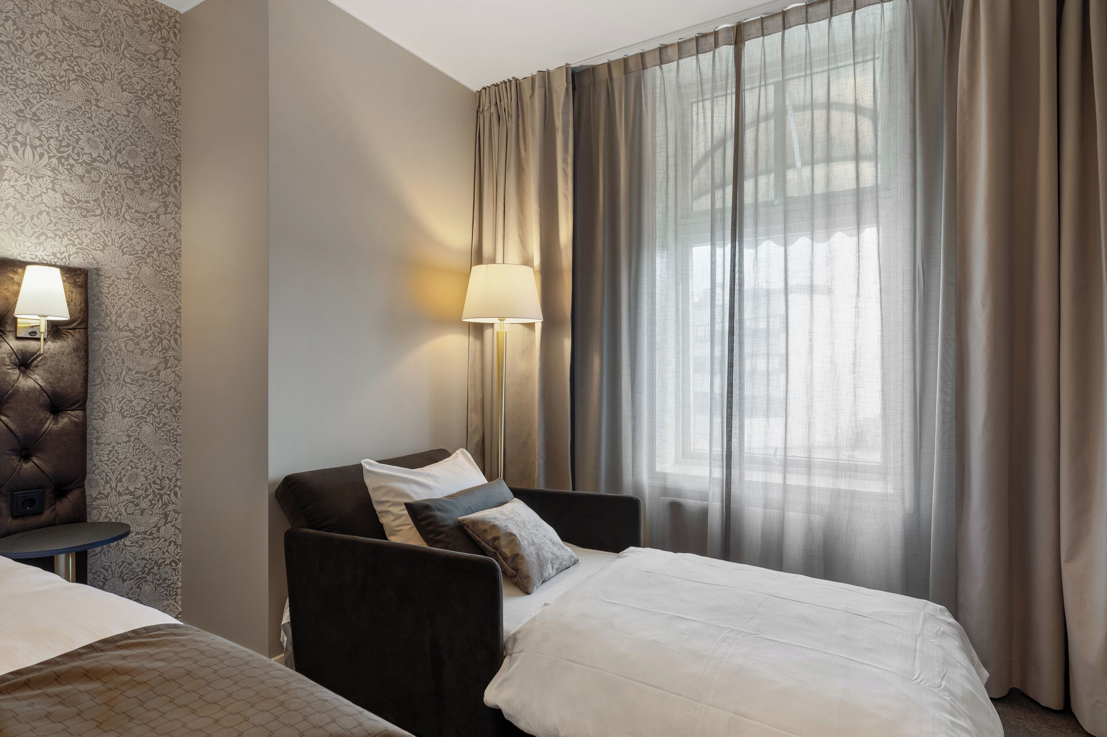 Habitación de hotel con dos camas, una mesita de noche, una lámpara y un ventanal con cortinas.