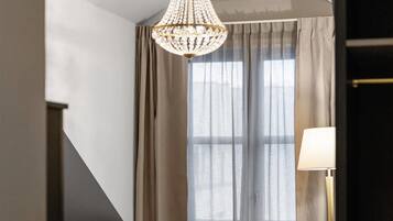 A hotel room with a bed, a chandelier, a lamp, a small table, and a window with curtains.