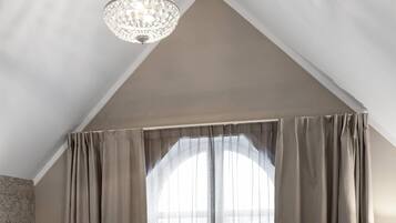 A bedroom with a bed, two chairs, a small table, a chandelier, and a window with curtains.