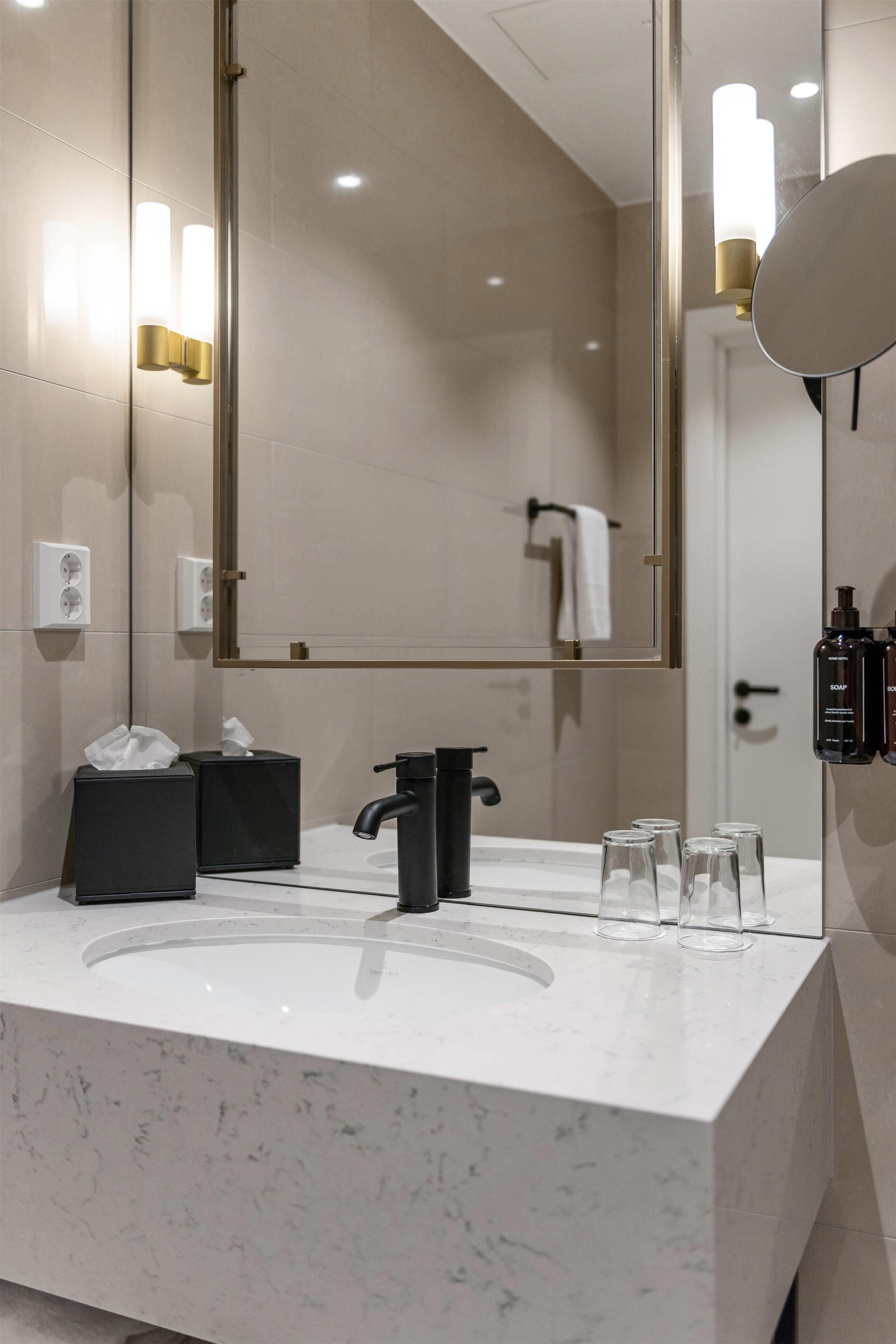 A modern bathroom with a large mirror, a white marble sink, black soap dispensers, and a towel rack.