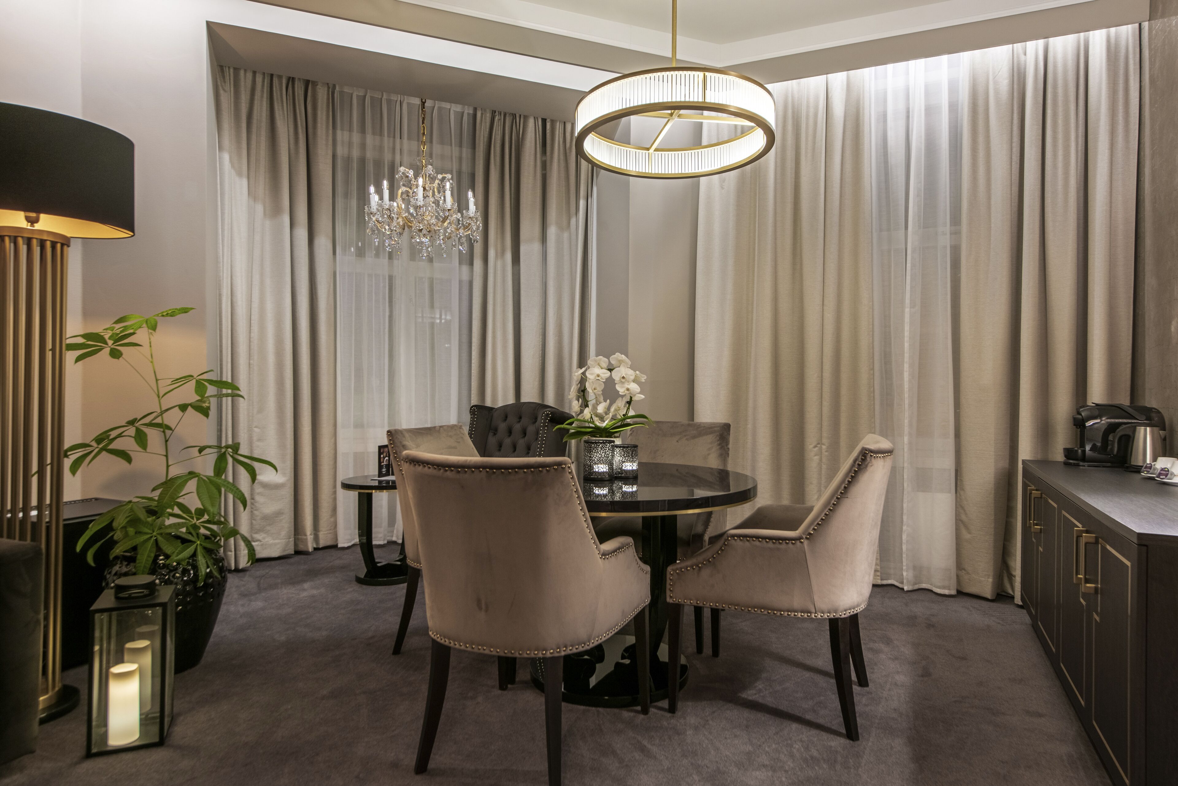 A dining area with a round table, four upholstered chairs, a chandelier, a potted plant, and a coffee machine.