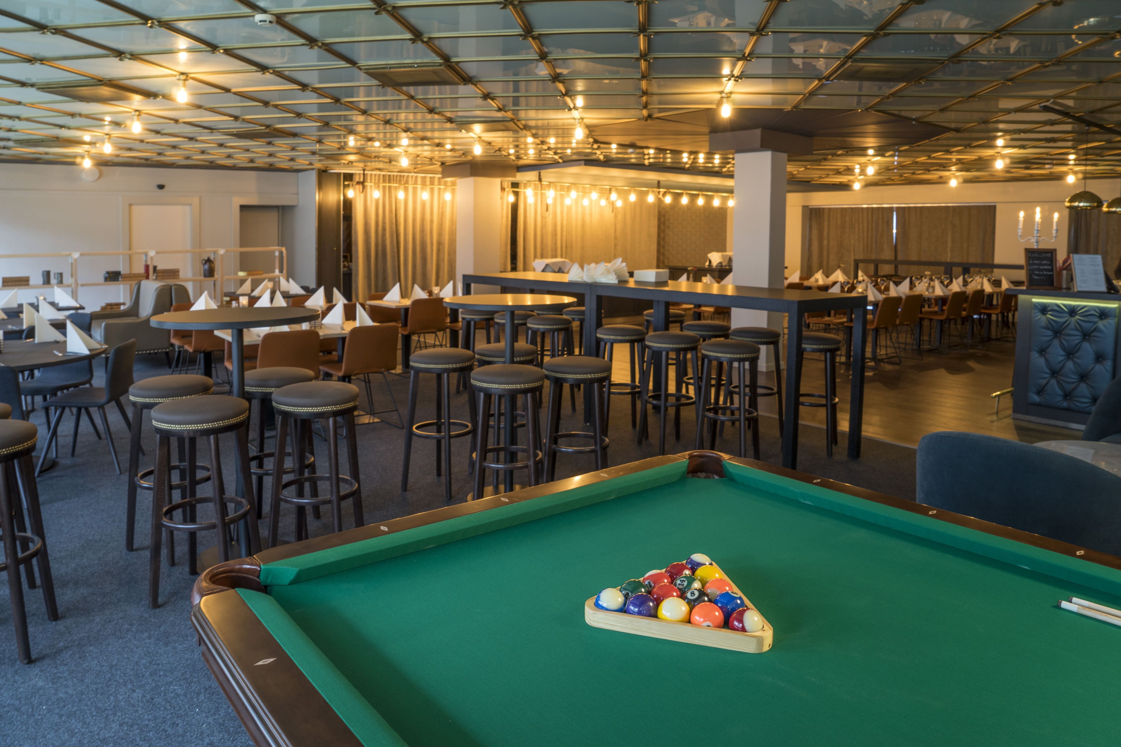 A pool table with balls, surrounded by bar stools and tables in a room with a high ceiling and pendant lighting.