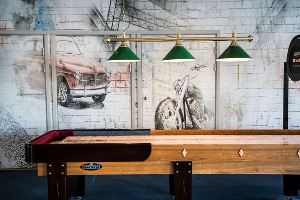 Una mesa de billar con tres lámparas verdes arriba, frente a una pared con un mural de coches antiguos y una motocicleta.