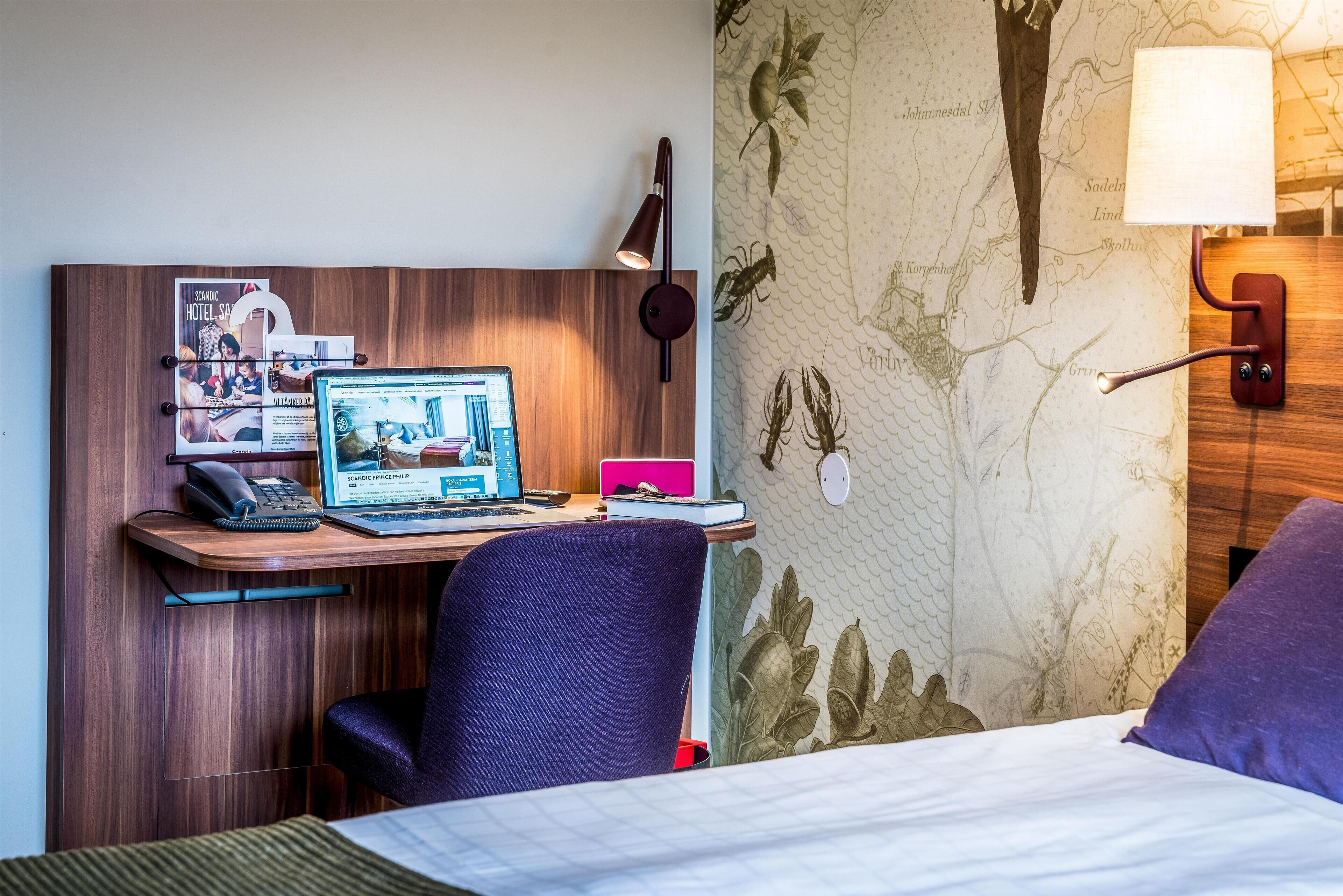A hotel room with a wooden desk, a laptop, a telephone, and a map on the wall.