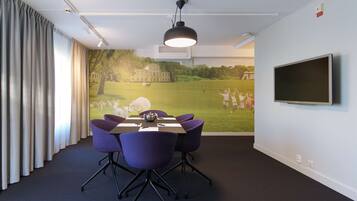 Un salón de conferencias con una mesa, sillas, una pantalla de proyector y un mural que muestra un campo con gente.