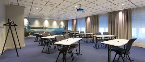 Un salón de conferencias con techo cuadriculado, varias mesas con sillas, un proyector y un mural decorativo en la pared.