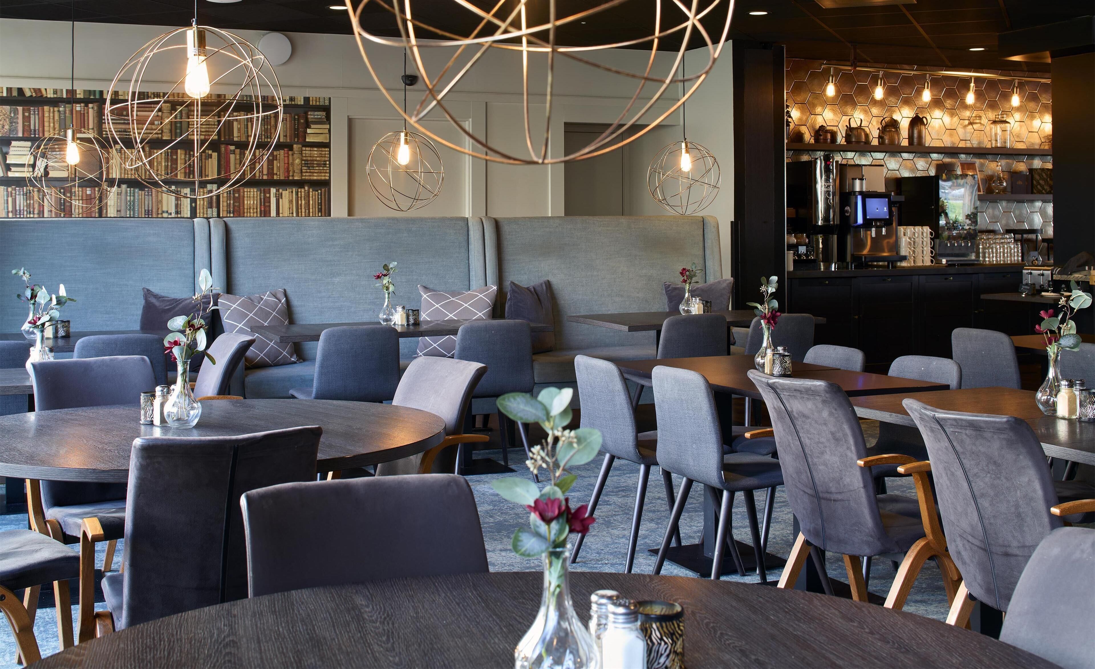 A modern restaurant interior with round tables, grey upholstered chairs, and decorative lighting.