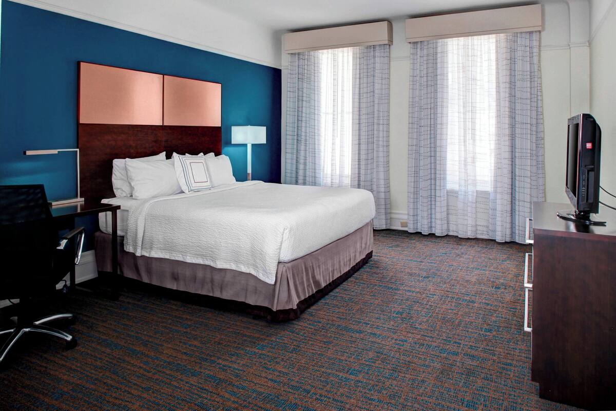 A hotel room with a large bed, a desk with a chair, a TV, and a window with curtains.