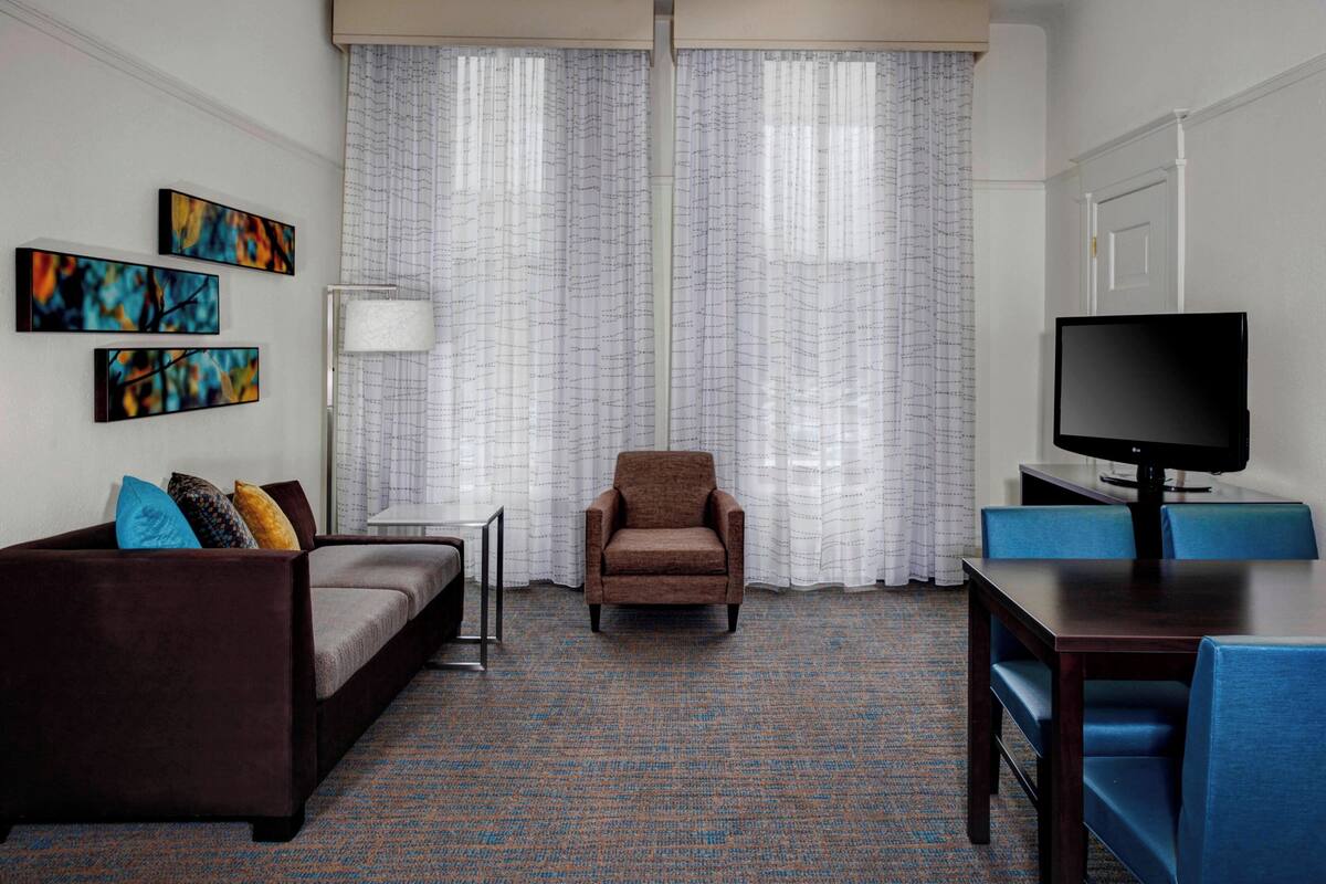 A hotel room with a brown sofa, a small armchair, a TV on a stand, and a dining table with blue chairs.