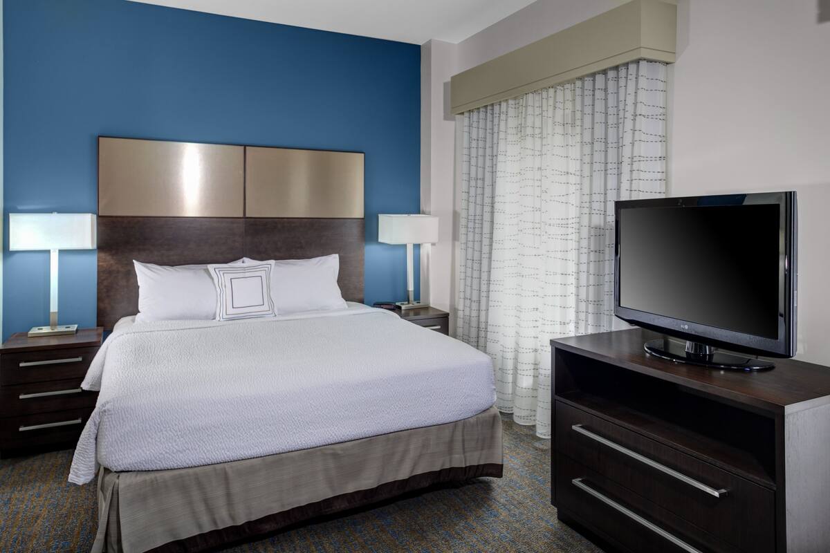A hotel room with a large bed, a flat-screen TV, a dresser, and a nightstand with lamps.