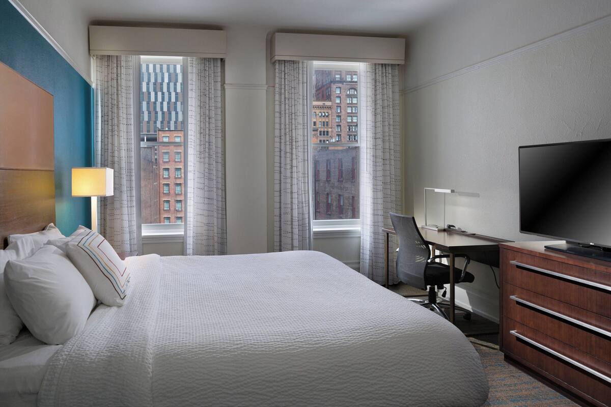 A hotel room with a large bed, a desk, a television, and a view of buildings through the windows.