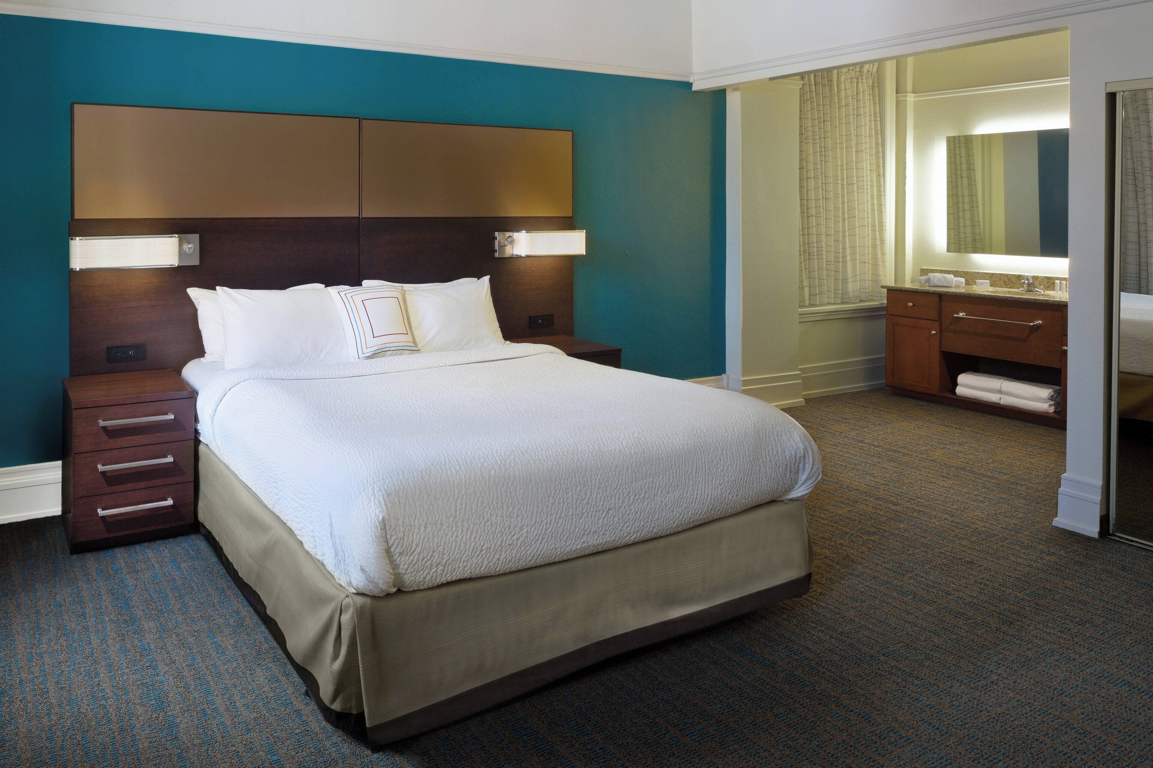 Quarto de hotel com uma cama grande, cabeceira de madeira, mesa de cabeceira, cômoda e banheiro com espelho e porta-toalhas.