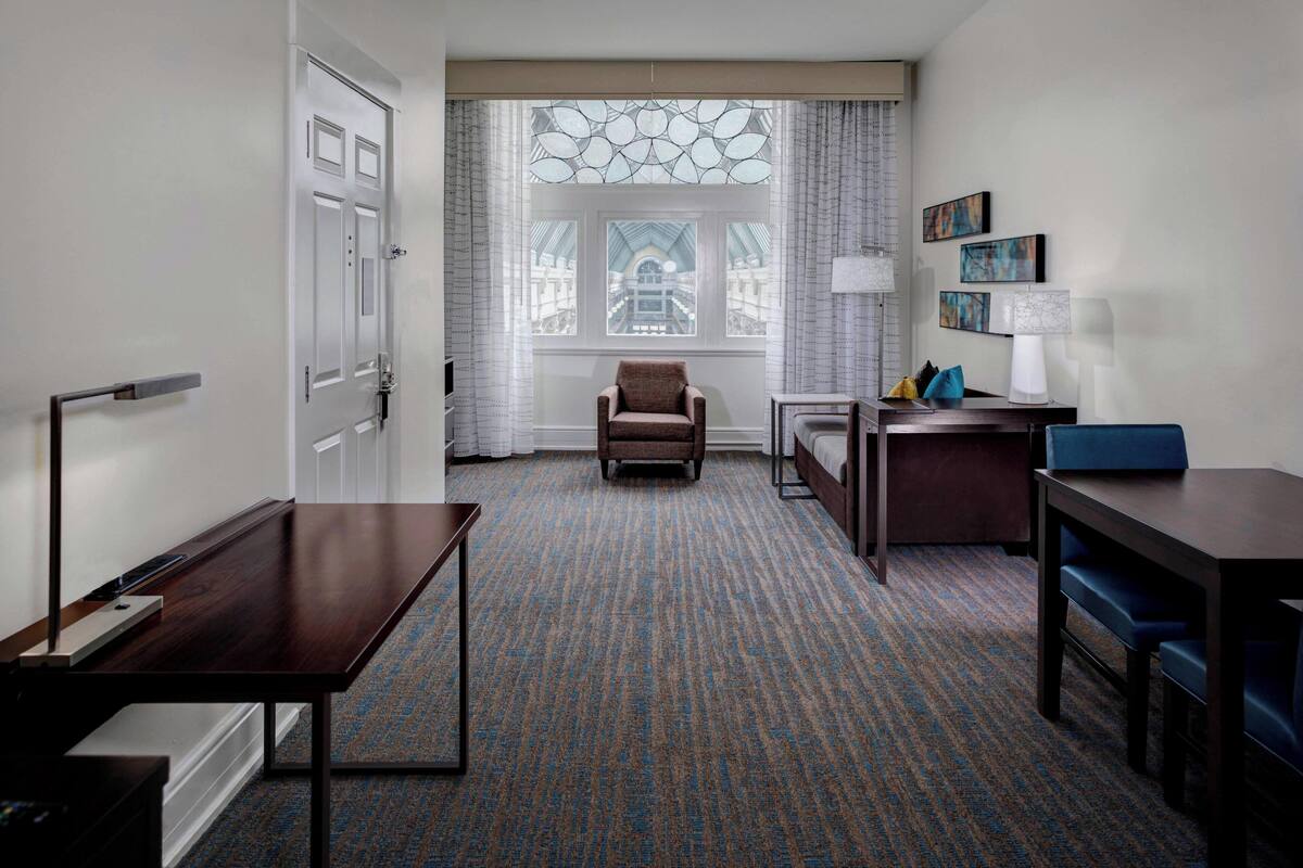 A hotel room with a large window, a chair, a desk, and a table.