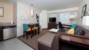 A compact hotel room with a kitchenette, a sofa, a dining area, and a TV.