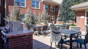 An outdoor patio with a grill, tables, and chairs, surrounded by brick buildings and evergreen trees. - Residence Inn by Marriott Boulder Broomfield (Louisville)