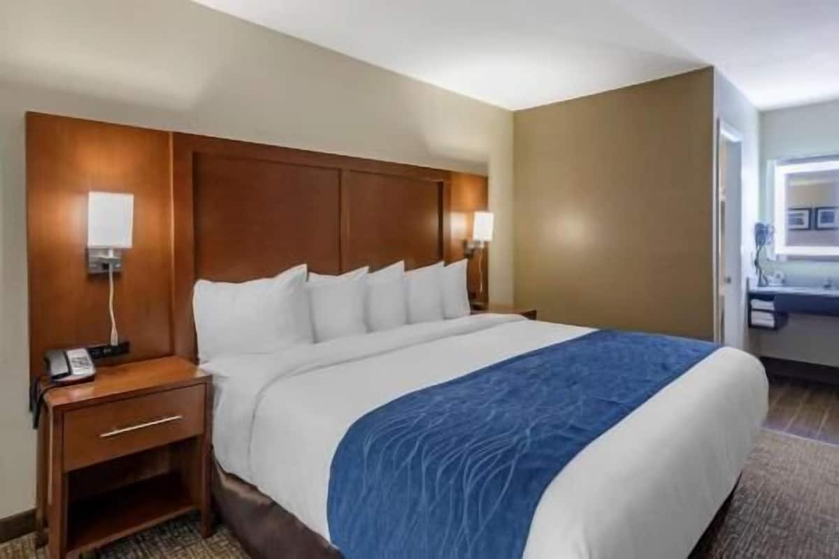 Une chambre d’hôtel avec un grand lit, une tête de lit en bois, deux tables de chevet avec des lampes et une vue sur le couloir.
