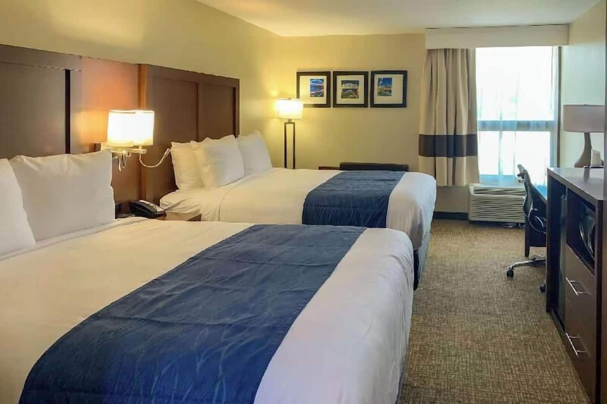 Une chambre d’hôtel avec deux lits, un bureau, une chaise, une lampe et une fenêtre avec des rideaux.
